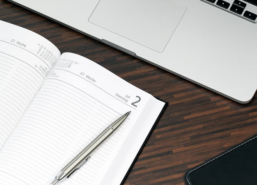 An open planner and a pen beside a laptop on a desk